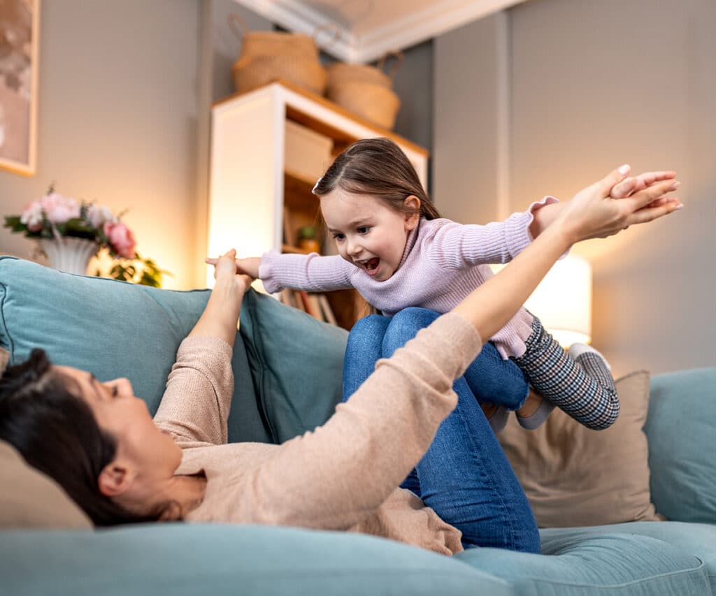 A mother lying on a teal sofa lifts her laughing young daughter into the air like an airplane in a warmly lit, cozy living room