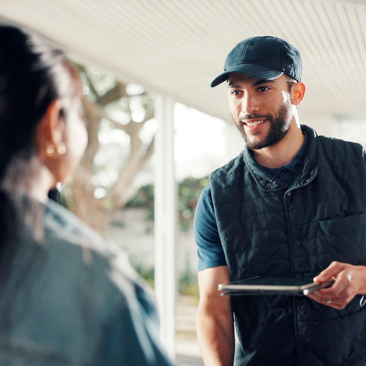 A smiling home service technician in a dark cap and vest holds a tablet while speaking with a female homeowner at her front door