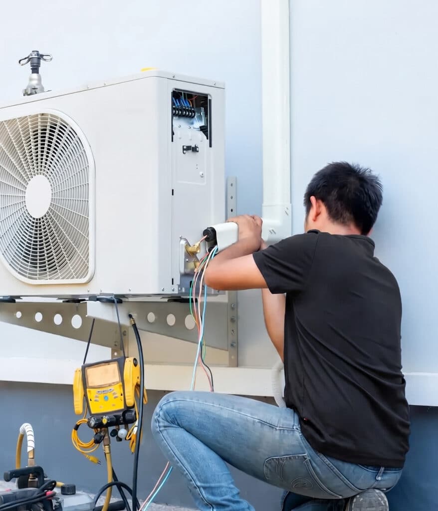 An HVAC technician in a black shirt crouches while connecting wiring on an outdoor air conditioning unit, with a yellow manifold gauge set on the ground nearby