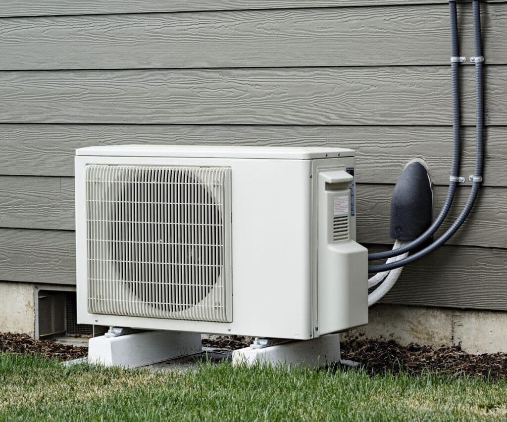 A white heat pump outdoor air conditioning unit mounted on concrete pads against a gray wood-grain siding house exterior, with refrigerant lines running up the wall